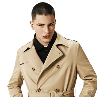 A man wearing a beige trench coat with buttons and belt at the waist, over a black shirt.